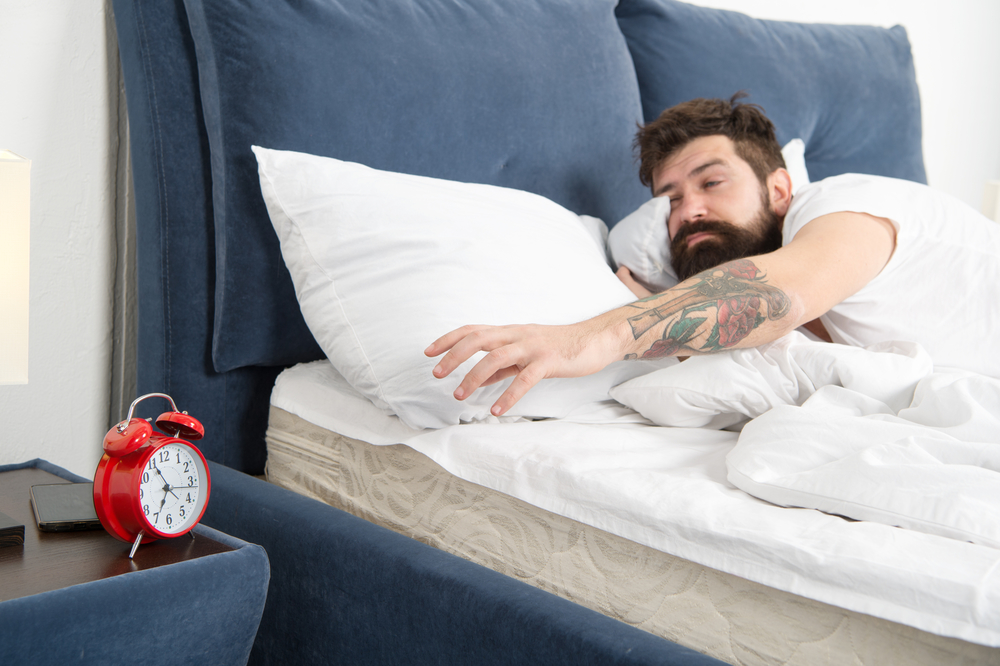 Overslept again. Tips for waking up early. Man bearded hipster sleepy face bed with alarm clock. Turn off that ringing. What terrible noise. Problem early morning awakening. Get up with alarm clock
